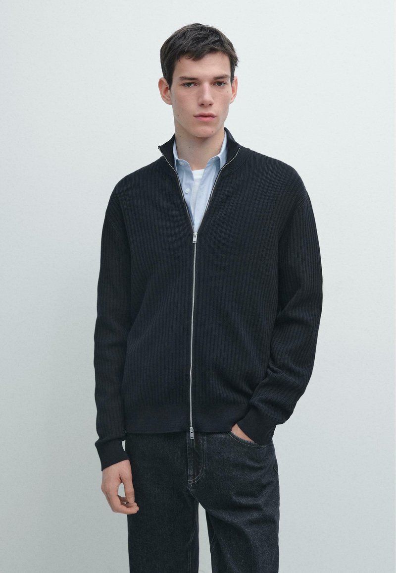 Schwarzer, gerippter Cardigan mit Reißverschluss und hohem Kragen aus Baumwollmischgewebe, lange Ärmel und eng anliegende Bündchen, getragen über einem hellblauen Hemd und dunkelblauen Jeans.