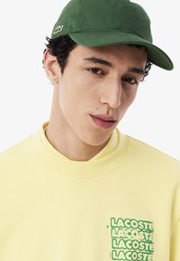 Groene katoenen pet gecombineerd met een gele sweatshirt die overlappende groene "LACOSTE"-tekst op de voorkant heeft. Zachte textuur, casual ontwerp.