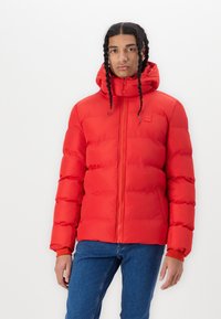 Rød pufjakke med hætte, der har vandret quilting, en frontlynlås og elastiske manchetter. Stylet med blå jeans.