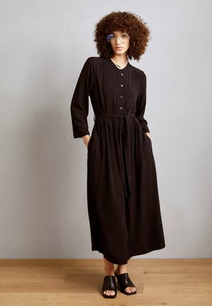 Robe noire à boutons, avec une taille cintrée, des manches longues, des poches latérales et un tissu doux. Portée avec des mules ouvertes noires.