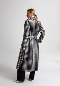 Długi płaszcz w wzór houndstooth; czarna i biała tkanina z paskiem. Posiada kołnierz, kieszenie z rozcięciem i długie rękawy. Noszony na czarnych spodniach.