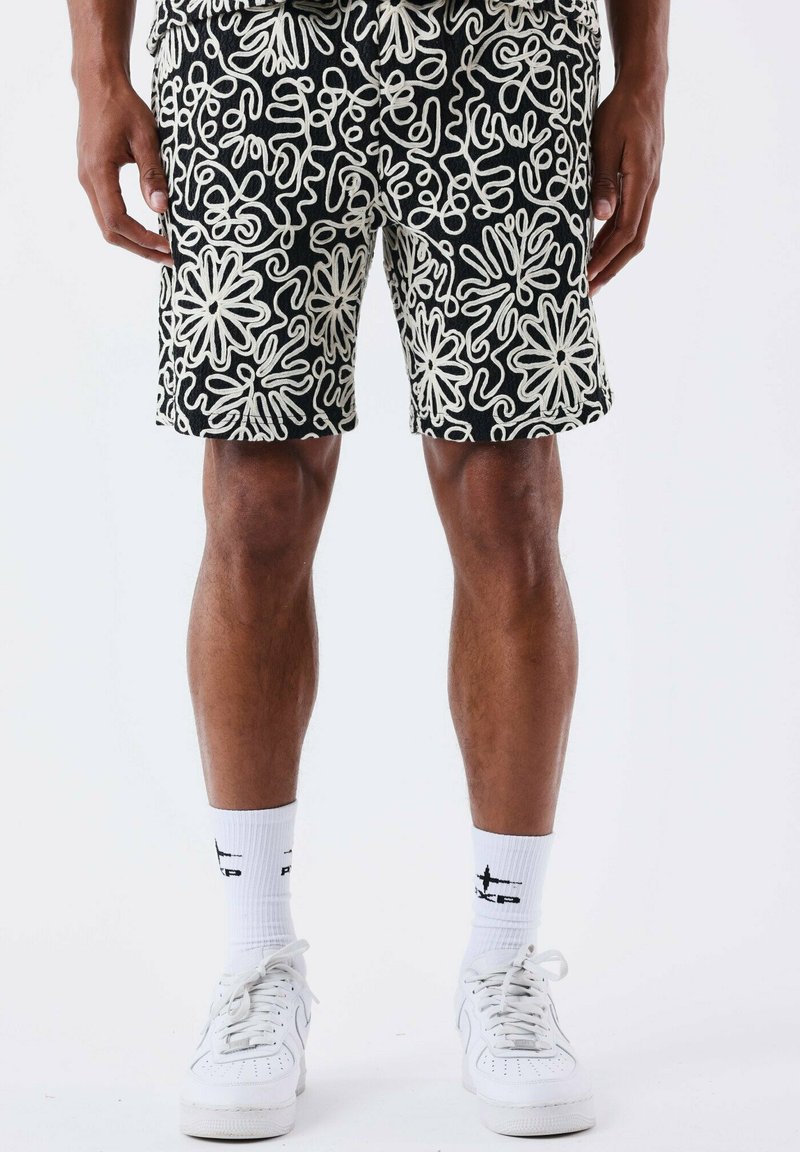 Shorts noirs avec des motifs floraux abstraits blancs, fabriqués en tissu texturé. Portés avec des chaussettes blanches et des baskets pour un look décontracté.