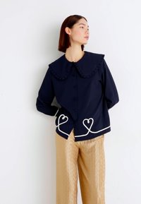 Ναυτικό μπλε πουκάμισο με κουμπιά και μεγάλο φουσκωτό γιακά, διακοσμημένο με λευκή καρό κουκίδα στο τελείωμα. Συνδυασμένο με ανοιχτόχρωμο, υφασμένο μπεζ παντελόνι.