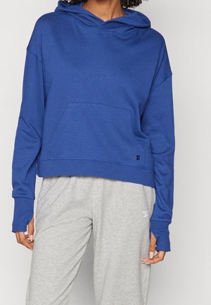 Kvinde iført en blå cropped hættetrøje med lange ærmer, frontlomme og lysegrå joggingbukser.