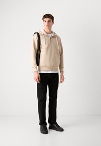 Haut à zip beige, t-shirt blanc en dessous, pantalon noir ajusté et baskets noires. Le modèle se tient avec un sac à bandoulière noir.