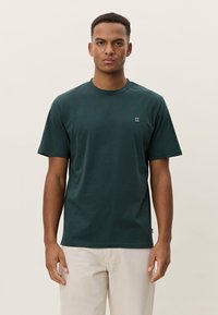 Σκούρο πράσινο βαμβακερό T-shirt με στρογγυλή λαιμόκοψη και κοντά μανίκια. Διαθέτει μικρό λευκό λογότυπο στο αριστερό στήθος. Απλός, ευθύγραμμος σχεδιασμός.