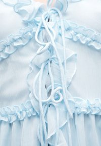 Υφασμα ανοιχτού μπλε χρώματος με βολάν στις άκρες και δεμένες κορδόνια, που παρουσιάζει λεπτή υφή και περίτεχνη λεπτομέρεια ενδύματος.