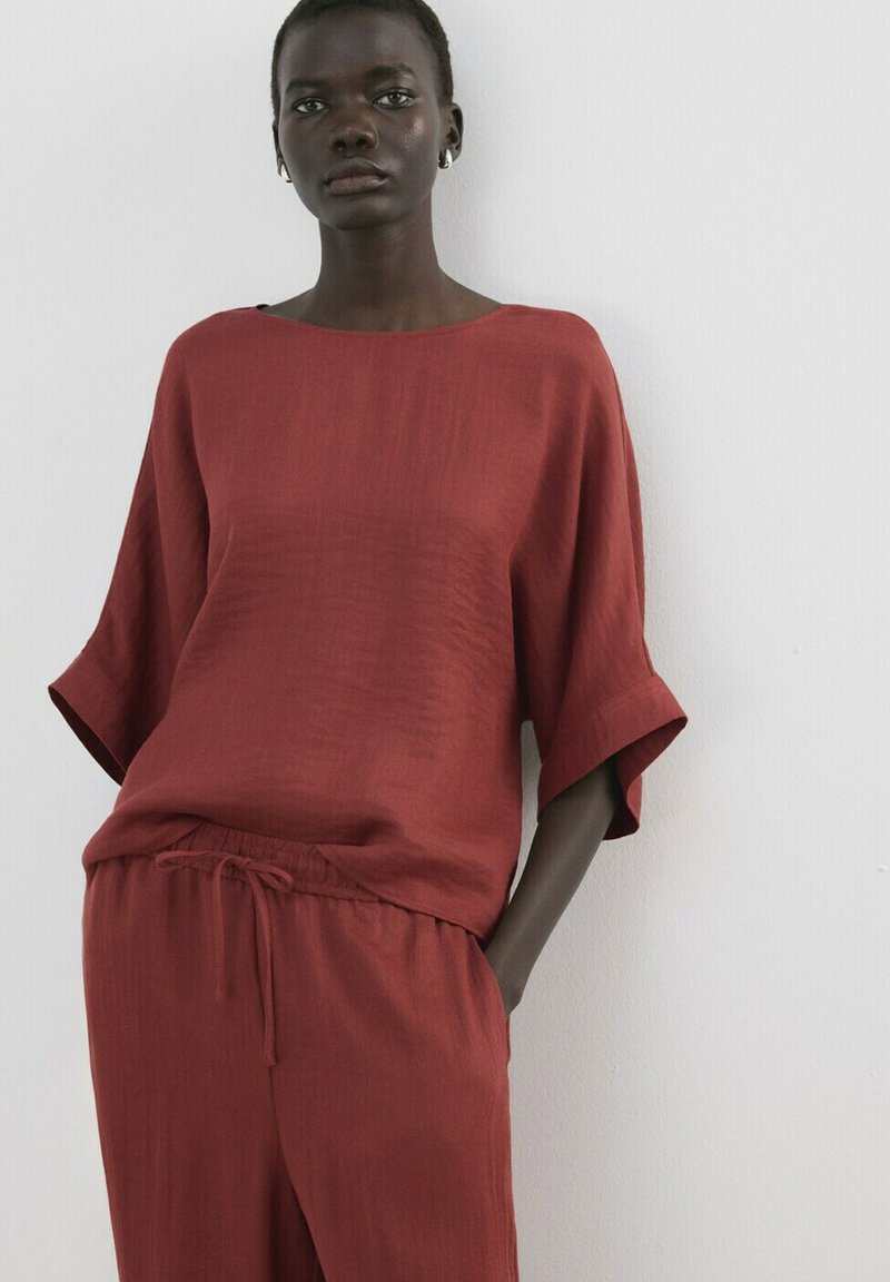 Femeie purtând un top larg cu mâneci scurte, de culoare roșu-ruginiu, și pantaloni cu șnur asortați, stând în fața unui perete alb simplu, cu o mână în buzunar.
