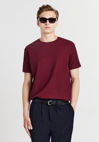 T-shirt bordeaux a maniche corte con collo rotondo, abbinata a pantaloni scuri e una cintura nera. Modello che indossa occhiali da sole rettangolari scuri.