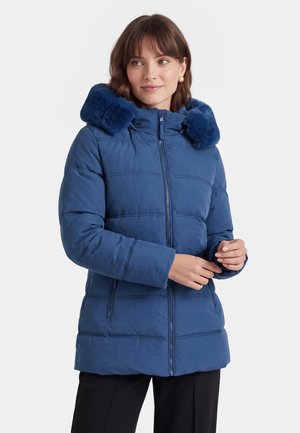 Donna che indossa una giacca invernale imbottita blu con cappuccio bordato in pelliccia, in piedi con le mani vicino alla cerniera su sfondo bianco.