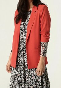 Blazer rosso realizzato in tessuto strutturato con una vestibilità su misura, abbinato a un vestito floreale nero con gonna arricciata.