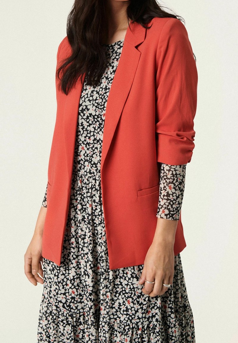 Blazer rosso realizzato in tessuto strutturato con una vestibilità su misura, abbinato a un vestito floreale nero con gonna arricciata.