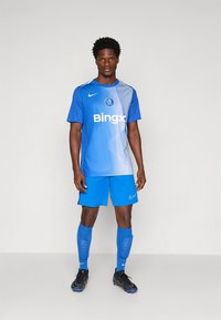 Maillot de football bleu avec un design en dégradé, logo blanc, et shorts bleus. Le joueur porte des crampons noirs et des chaussettes bleues. Texture de tissu lisse.