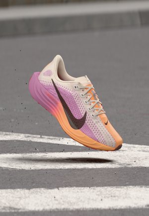 Lengvas bėgimo batelis su rožinės, oranžinės ir smėlio spalvų gradientiniu mezginiu, juodu Nike logotipu, ore virš grublėto asfalto su baltomis kelio žymėmis.