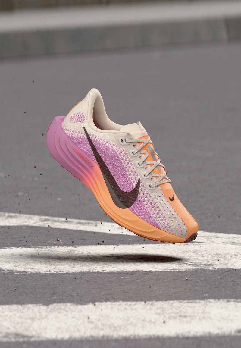 Ελαφρύ παπούτσι για τρέξιμο με ροζ, πορτοκαλί και μπεζ βαθμιαίο πλεκτό, μαύρο σήμα Nike, ακουμπάει στον αέρα πάνω από τραχιά άσφαλτο με λευκές οδικές γραμμές.