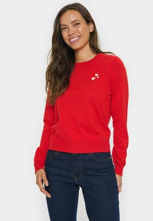 Rood gebreid sweatshirt met lange mouwen, ronde halslijn en drie kleine witte hartapplicaties op de borst. Gedragen met donkerblauwe jeans.