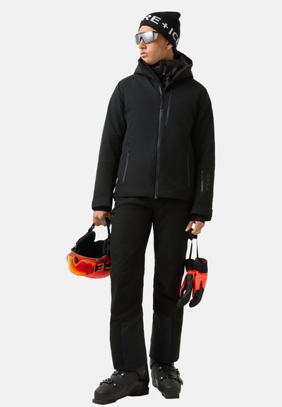 Sort skijakke med hætte, sorte bukser, der holder en rød skihjelm og orange goggles. Har tekstureret materiale og lynlåse.