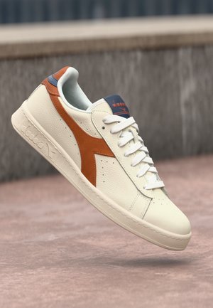 Cream leather sneaker with orange and navy accents, rounded toe, flat white laces, textured sole, and branding on tongue and side.