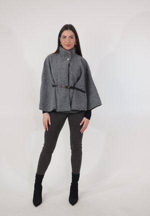 Frau steht und trägt einen grauen Wollcape mit Gürtel, schwarze eng anliegende Hosen und schwarze Stiefeletten vor weißem Hintergrund.