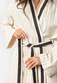 Bež bathrobe sa crnim detaljima, širokim rukavima i vezanim pojasom. Karakteristike uključuju meku teksturu i dva prednja dzepa.