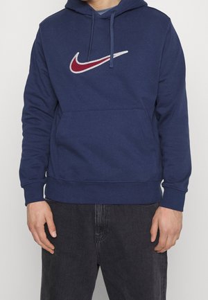 Osoba ubrana w granatową bluzę z kapturem z czerwonym i białym logo Nike w kształcie łyżwy, w zestawie z czarnymi dżinsami, stojąca na jednolitym tle.