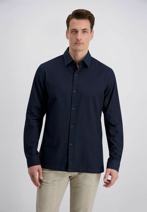 Man draagt een donkerblauw overhemd met lange mouwen en lichte beige broek, staand tegen een effen lichtgrijze achtergrond.