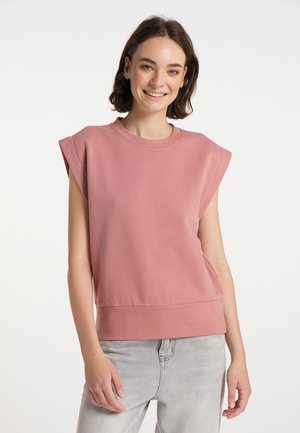 Young woman smiling, wearing sleeveless dusty pink top and light gray jeans, standing against a plain white background.