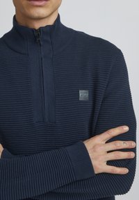 Marineblauer, gerippter Strickpullover mit einem halben Reißverschluss-Kragen und einem kleinen Logo-Patch auf der Brust. Verfügt über strukturierte Ärmel und Bündchen.