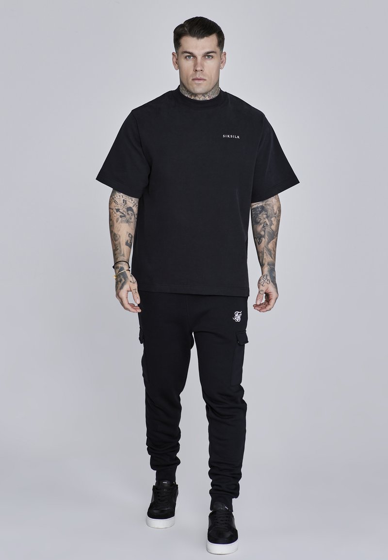 Crna majica s okruglim izrezom i kratkim rukavima, u kombinaciji s crnim cargo trenirkama s bočnim džepovima i bijelim logotipom na bokovima.