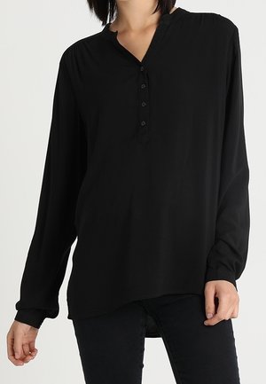 Vrouw draagt een loszittende zwarte blouse met lange mouwen en een knoopsluiting, gecombineerd met een zwarte broek, tegen een witte achtergrond.