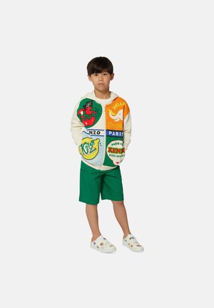Veste à zip colorée avec des graphismes variés, corps crème et accents verts. Associée à un short vert et des chaussures blanches avec des motifs colorés.