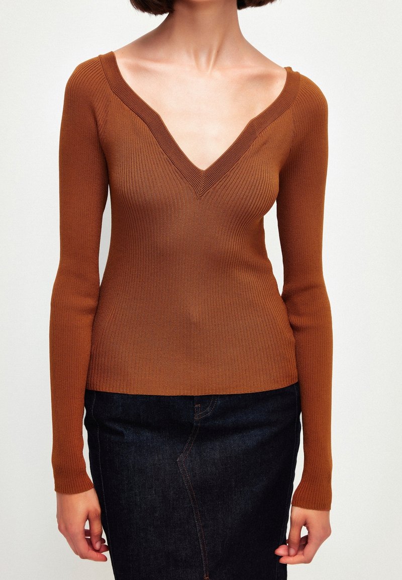 Femme portant un pull marron moulant à manches longues, à col en V côtelé, associé à une jupe en jean foncé sur un fond uni.