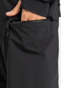 Zwarte softshell broek met een ritszak aan de zijkant. Het materiaal is soepel en rekbaar, wat zorgt voor gemak in beweging.