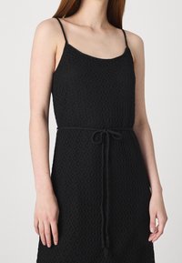 Černé bezrukávové šaty s texturovaným tkaným vzorem, vybavené pásem na stažení pro úpravu a uvolněným střihem.