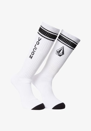 Calcetines blancos hasta la rodilla con acentos negros, con textura acanalada, dos rayas negras en la parte superior y un logo en el lateral.