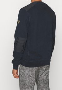 Marinblå sweatshirt med rund halsringning, vadderade axelpatchar och ribbade ärmslut. Bärs med grå, texturerade sweatpants.