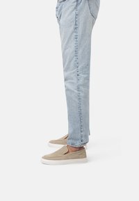 Zapatillas de ante beige tipo slip-on con suela de goma blanca, que presentan una textura suave y un diseño minimalista. Combinadas con pantalones de mezclilla de color azul claro.