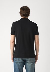 Svart polo skjorte med korte ermer, ribbestrikket krage og mansjetter, laget av glatt stoff, brukt med blå denim jeans. Vist fra baksiden.
