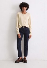 Beige gestrickte Strickjacke mit Rundhalsausschnitt und Knopfverschluss, kombiniert mit dunkelblauen Jeans im geraden Schnitt und braunen Schnürschuhen.