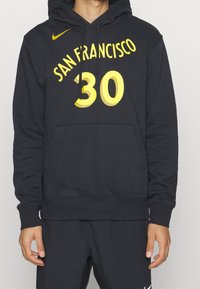 Personne portant un hoodie noir avec le texte jaune "San Francisco 30" et le logo Nike, associé à un short noir avec le swoosh blanc de Nike.
