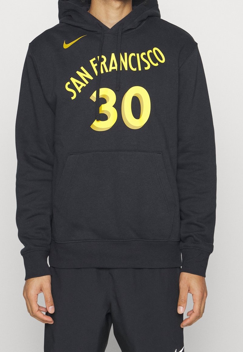 Personne portant un hoodie noir avec le texte jaune "San Francisco 30" et le logo Nike, associé à un short noir avec le swoosh blanc de Nike.