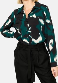 Woman wearing a teal, black, and white abstract patterned button-up blouse tucked into high-waisted black pants with a tied belt.