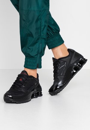Persoon die groene joggingsbroek en zwarte Nike Shox sneakers met zichtbare demping en veters draagt, staand op een witte vloer.