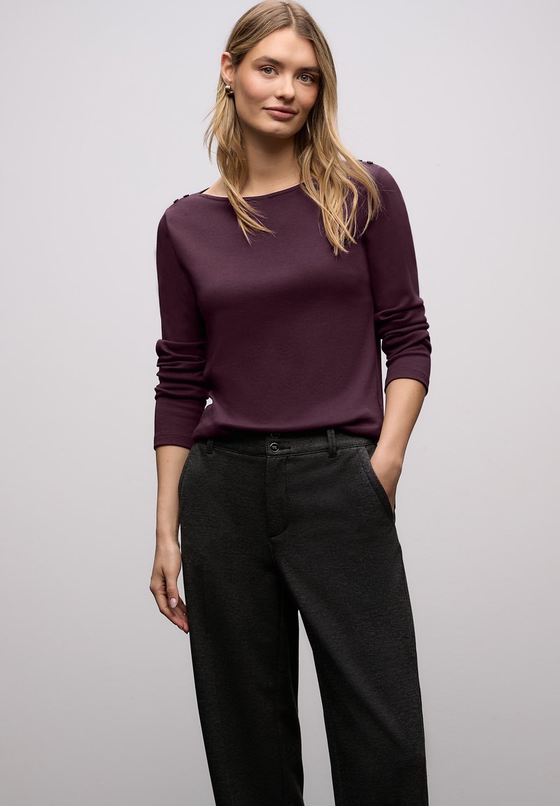 Giovane donna con i capelli biondi lunghi, indossa una maglia a maniche lunghe viola scuro e pantaloni grigio scuro, in piedi davanti a uno sfondo chiaro uniforme.