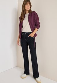 Giacca zip-up bordeaux, top a maglia beige, jeans neri a palazzo con cuciture in contrasto e sneaker bianche con suola beige.