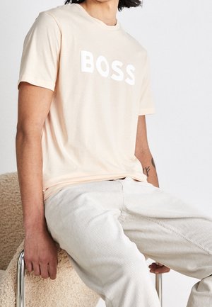 Persoon draagt een lichtbeige t-shirt met de tekst "BOSS" en lichtgekleurde broek, zittend op een beige gestructureerde stoel met metalen poten.