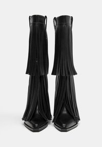 Črne usnjene gležnjarje z dolgimi frinži, koničastim nosom in masivnim peto. Teksturirani frinži dodajajo dinamično gibanje.