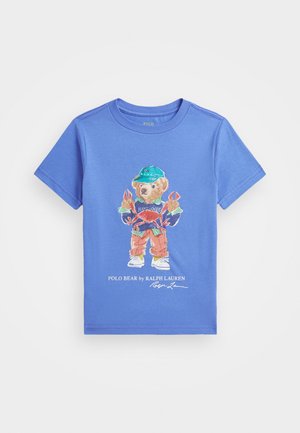 Blauw kinder T-shirt met een beer die een groene pet, gestreept shirt, oranje broek draagt, een rode krab vasthoudt en witte sneakers draagt.
