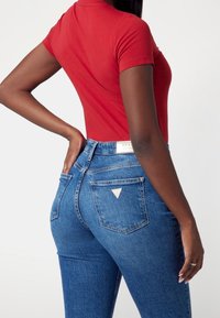 Červené přiléhavé tričko v kombinaci s modrými džínami s vysokým pasem. Džíny mají zadní kapsy a trojúhelníkovou značkovou etiketu na pase.