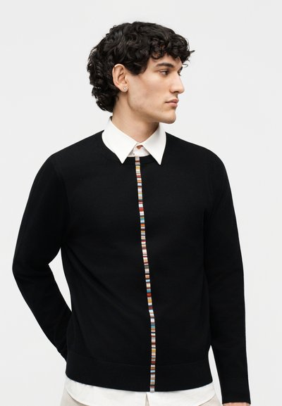 Νεαρός με κατσαρά μαλλιά, που φοράει μαύρο πουλόβερ με πολύχρωμη κάθετη ρίγα και λευκό πουκάμισο με γιακά από κάτω.